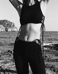 Person wearing a black bikini top and pants standing on rocky terrain by the sea.